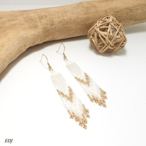 Peut inclure: Boucles d'oreilles pendantes en perles blanches et dorées avec un design en frange. Les boucles d'oreilles sont faites de petites perles rondes et ont une fermeture à crochet dorée.