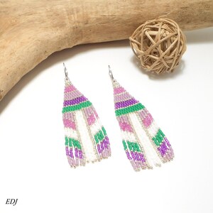 May include: A pair of beaded earrings with a feather design. The earrings feature a combination of white, pink, green, and purple beads. The earrings have a silver-colored hook closure. The earrings are on a white surface.