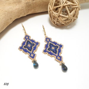 May include: A pair of gold dangle earrings with a blue and gold geometric pattern and a dark blue teardrop-shaped stone hanging from each earring.