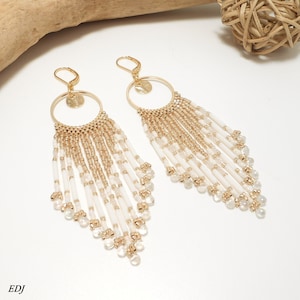 May include: A pair of long, gold hoop earrings with white and gold beads hanging from them. The earrings are in a teardrop shape and have a delicate, bohemian style.