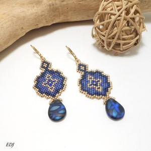 May include: A pair of blue and gold beaded earrings with a teardrop-shaped blue stone pendant. The earrings are made with small beads and have a geometric design.