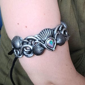 Op de afbeelding: Zilverkleurige armmanchet met een schelpmotief en een iriserende edelsteen in het midden. De metalen manchet heeft een zwarte koordsluiting. De armsieraad is een uniek accessoire.