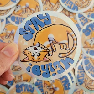Peut inclure: Un autocollant rond avec un chat orange de dessin animé et les mots "STAY WEIRD" en bleu. Le chat est à l'envers. L'autocollant a un effet holographique et est entouré d'autres autocollants similaires.