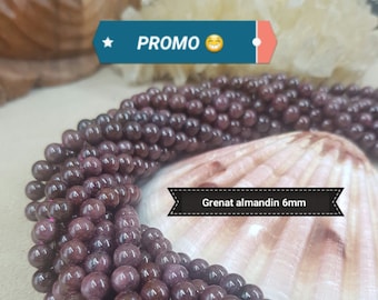 May include: A strand of dark red gemstone beads, likely garnet, with a diameter of 6mm. The beads are arranged in a circular pattern and are displayed on a white seashell. The text "Grenat almandin 6mm" is visible in the image.