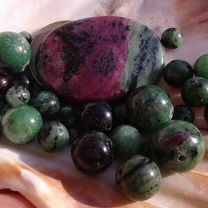 Peut inclure: Un ensemble de perles de pierres précieuses vertes, noires et roses et une pierre précieuse plus grande de forme ovale, verte, noire et rose. Les pierres précieuses sont disposées sur une surface blanche texturée.