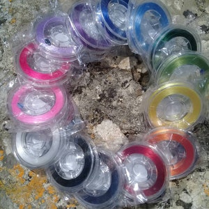 Può includere: Una collezione di bobine di filo colorato, avvolte in plastica trasparente, disposte in cerchio su una superficie di pietra. I colori del filo includono rosa, viola, blu, verde, giallo, arancione, rosso, nero e bianco.