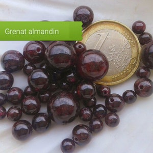 Può includere: Primo piano di un mucchio di perle di granato rosso scuro, alcune delle quali sono rotonde e altre leggermente irregolari. Le perle sono sparse su una superficie bianca, e una moneta da 1 euro è visibile sullo sfondo. Il testo "Grenat almandin" è visibile nell'immagine.