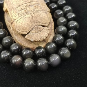 Può includere: Un braccialetto di perline di ossidiana nera con un ciondolo a forma di scarabeo in legno intagliato. Le perline sono rotonde e hanno una finitura lucida.