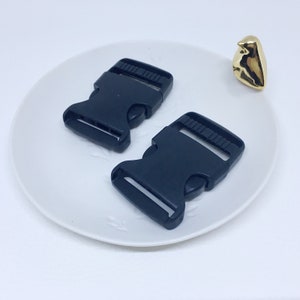 May include: Two black plastic side release buckles on a white plate. The buckles are approximately 5 cm long and 2.5 cm wide.