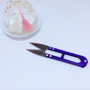 Peut inclure: Ciseaux à fils violets avec une lame argentée et un dôme en verre transparent contenant deux pompons roses et blancs à l'intérieur.