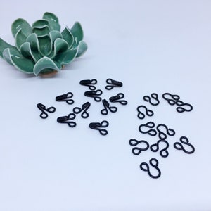 Peut inclure: Une collection de fermoirs à crochet et à œil en métal noir éparpillés sur une surface blanche. Les crochets et les yeux sont utilisés pour la couture et l'artisanat.