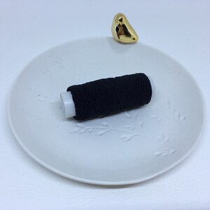 May include: A spool of black thread on a white ceramic plate with a floral design. A small, gold-colored bird figurine sits on the plate's edge. The thread is tightly wound around a white plastic core.