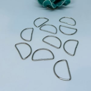 May include: Ten silver-toned metal D-rings, also known as D rings or D-shaped rings, are scattered on a white surface. These rings are commonly used in crafting and sewing projects.