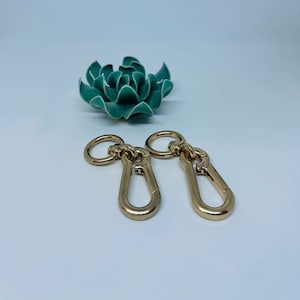 May include: Two gold-colored metal clasps with a ring on each end. The clasps are open and ready to be attached to a bag or other item.
