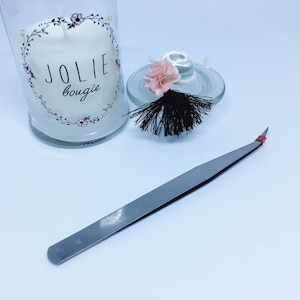 May include: A silver pair of tweezers with a red tip, a glass jar with a white candle and a black tassel, and a pink flower on top of the lid. The candle jar has a floral design and the text "Jolie bougie" on the front.