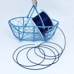 Puede incluir: Una bobina de cordón de algodón encerado azul oscuro en una cesta de alambre azul claro. El cordón se está desenrollando de la bobina y se encuentra sobre una superficie blanca.