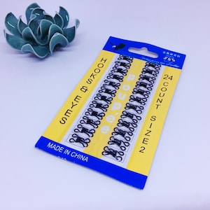 Peut inclure: Un paquet de crochets et d'yeux noirs, taille 2, avec 24 pièces. L'emballage est jaune et bleu, avec les mots "HOOKS & EYES" imprimés verticalement. Une succulente en céramique est en arrière-plan.