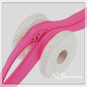 May include: Close-up of a bright pink zipper with a matching pull tab, partially unzipped. The zipper is wrapped around two white wooden spools. The text "NLB mercerie" is in the bottom right corner.