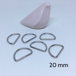 Peut inclure: Sept anneaux en D en métal argenté, chacun mesurant 20 mm de diamètre. Les anneaux sont disposés en demi-cercle sur une surface blanche.