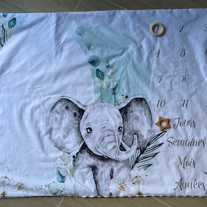 May include: A white baby milestone blanket with a black and white elephant illustration, green leaves, and white flowers. The blanket has numbers 1 through 12 printed on it, along with the words "Jours", "Semaines", "Mois", and "Années".