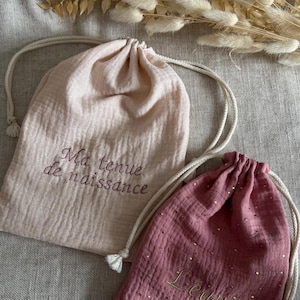 Op de afbeelding: Twee trekkoordzakken, één lichtroze en één mauve, met witte trekkoorden. De lichtroze tas heeft geborduurde tekst die luidt "Ma tenue de naissance". De mauve tas heeft gouden versieringen en geborduurde tekst "Leane".