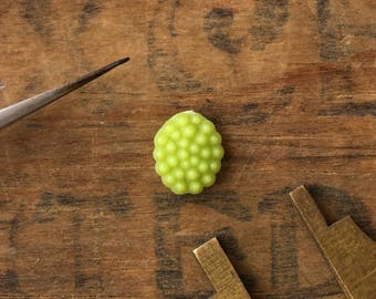 May include: A small, green, oval-shaped button with a textured surface. The button is made of plastic and has a raised, bumpy design.