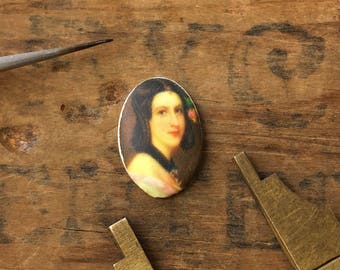 May include: Oval cameo featuring a portrait of a woman with dark hair and a white dress. The cameo is set in a silver frame and has a white background.