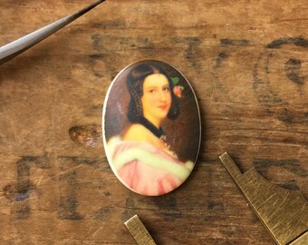 May include: Oval shaped cameo with a portrait of a woman with dark hair and a pink dress. She is wearing a flower in her hair. The cameo is white with a gold trim.