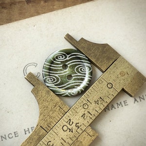 May include: A green button with a white swirl pattern is being measured with a brass ruler. The ruler has markings in inches.
