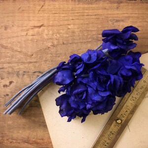 May include: A bunch of deep purple silk flowers with a gray stem, arranged on a vintage postcard with a brass ruler.