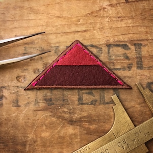 May include: A triangular patch with a brown felt base and a red felt top. The patch is sewn with pink thread.