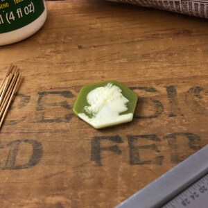 May include: A green hexagonal cameo with a white cameo of a woman's profile. The cameo is on a wooden surface.