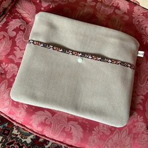 Peut inclure: Une pochette pour ordinateur portable en velours côtelé beige avec une bordure à motif noir et rose. La pochette a une fermeture à bouton unique et le mot "and" est imprimé sur le côté.