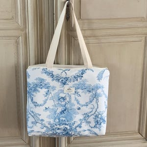 Peut inclure: Un sac cabas blanc avec un motif toile bleu et blanc représentant un dessin floral et un petit oiseau. Le sac a une fermeture éclair et deux longues anses.