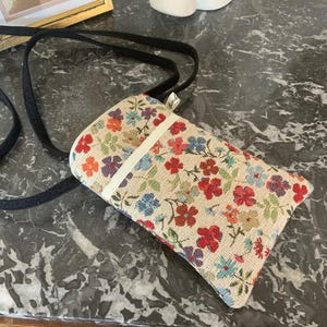 Peut inclure: Un étui pour téléphone à imprimé floral avec une sangle noire. L'étui présente un fond beige avec des fleurs rouges, bleues et orange. Une bande blanche traverse le centre de l'étui.