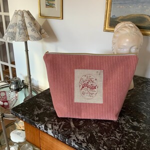 Könnte beinhalten: Eine rosa Kosmetiktasche aus Cord mit einem rechteckigen Aufnäher mit Text und einem ovalen Design. Die Tasche hat einen Reißverschluss und steht auf einer dunklen Marmoroberfläche.