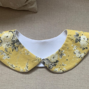 Peut inclure: Un col floral jaune avec un intérieur blanc texturé. Le col présente un motif de fleurs blanches et grises sur un fond jaune. Le col est conçu pour être porté autour du cou.