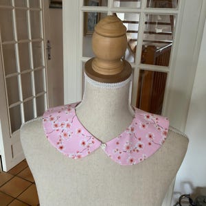 Peut inclure: Un col en tissu rose avec un bouton blanc et un imprimé floral de fleurs de cerisier roses sur fond blanc.