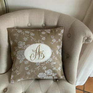Könnte beinhalten: Ein dekoratives Kissen mit einem floralen Muster in Beige und Weiß, mit einem gestickten Monogramm in einem Oval. Das Kissen liegt auf einem beige gepolsterten Stuhl, passend für die Inneneinrichtung.