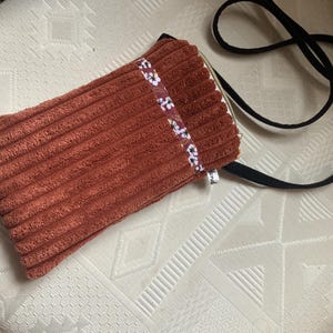 Peut inclure: Un sac bandoulière en velours côtelé couleur rouille avec une sangle noire. Le sac présente un ruban à motifs floraux et une fermeture éclair. Le sac est rectangulaire et a un aspect doux et texturé.