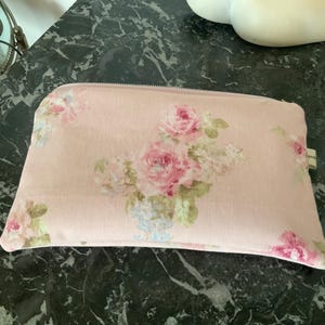Puede incluir: Una bolsa rectangular rosa claro con cremallera y un estampado floral. El estampado presenta rosas rosas, flores blancas y detalles azules. La bolsa es probablemente de tela.