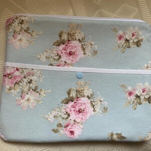 Peut inclure: Une housse pour ordinateur portable bleu clair avec un motif floral composé de roses roses et de feuilles vertes. La housse est dotée d'une fermeture éclair blanche et d'un bouton bleu clair.