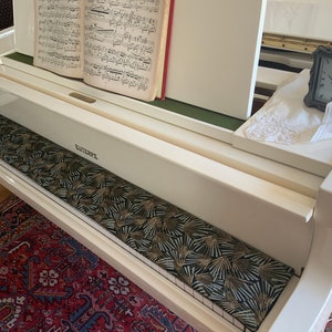 Può includere: Un pianoforte bianco con spartiti aperti sul leggio. Un cuscino decorativo per la panca con un motivo a ventaglio nero e oro si trova sopra i tasti. Un piccolo orologio e un fazzoletto di pizzo bianco sono sulla superficie del pianoforte. La parola "EUTERPE" è stampata sul pianoforte.