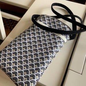 Peut inclure: Une housse de téléphone à motifs noirs et blancs avec une sangle noire. L'étui présente un motif répétitif en forme d'éventail avec des accents dorés. La sangle est un cordon fin et noir.