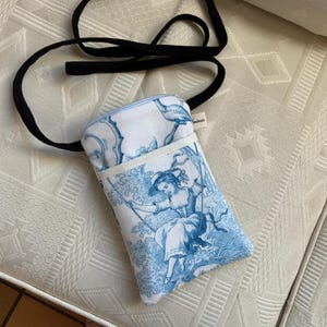 Peut inclure: Un sac de téléphone à imprimé toile blanc et bleu avec une sangle noire. Le sac présente une scène de toile classique d'une femme sur une balançoire. Le sac a une petite poche à l'avant et une fermeture éclair.