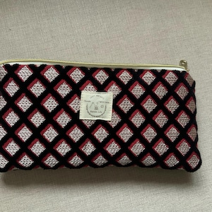 Può includere: Un astuccio rettangolare con cerniera e un motivo a rombi neri, rossi e argentati. L'astuccio ha una cerniera dorata e una piccola etichetta bianca con testo. Adatto per cosmetici o piccoli oggetti.