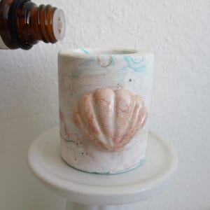 May include: A cylindrical concrete vessel featuring a marbled white, pink, and blue finish with a prominent, raised peach-colored seashell relief on the front. The container has an open top and a smooth, flat base, showcasing a textured surface.
