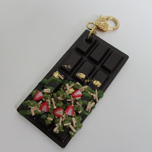 May include: A dark brown chocolate bar keychain featuring a gold-tone metal lobster clasp. The bar is decorated with green textured accents, small red and white strawberry slices, thin beige sticks, and scattered gold leaf flakes on the surface.