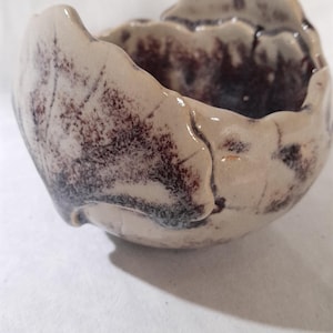 Peut inclure: Un petit bol en céramique fait à la main, de forme organique. Le bol présente un extérieur crème avec des motifs mouchetés brun foncé et noirs. L'intérieur est brun foncé. Le bord a un bord irrégulier et texturé.