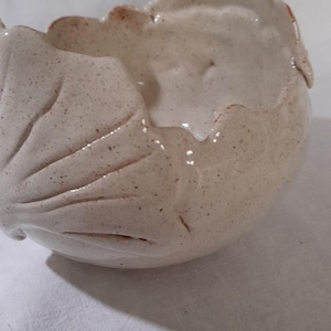 Peut inclure: Un bol en céramique fait à la main avec un motif de feuille. Le bol est blanc cassé avec des mouchetures marron et une surface texturée. Le bord a un bord ondulé avec des accents marron. Le bol mesure environ 15 cm de diamètre.
